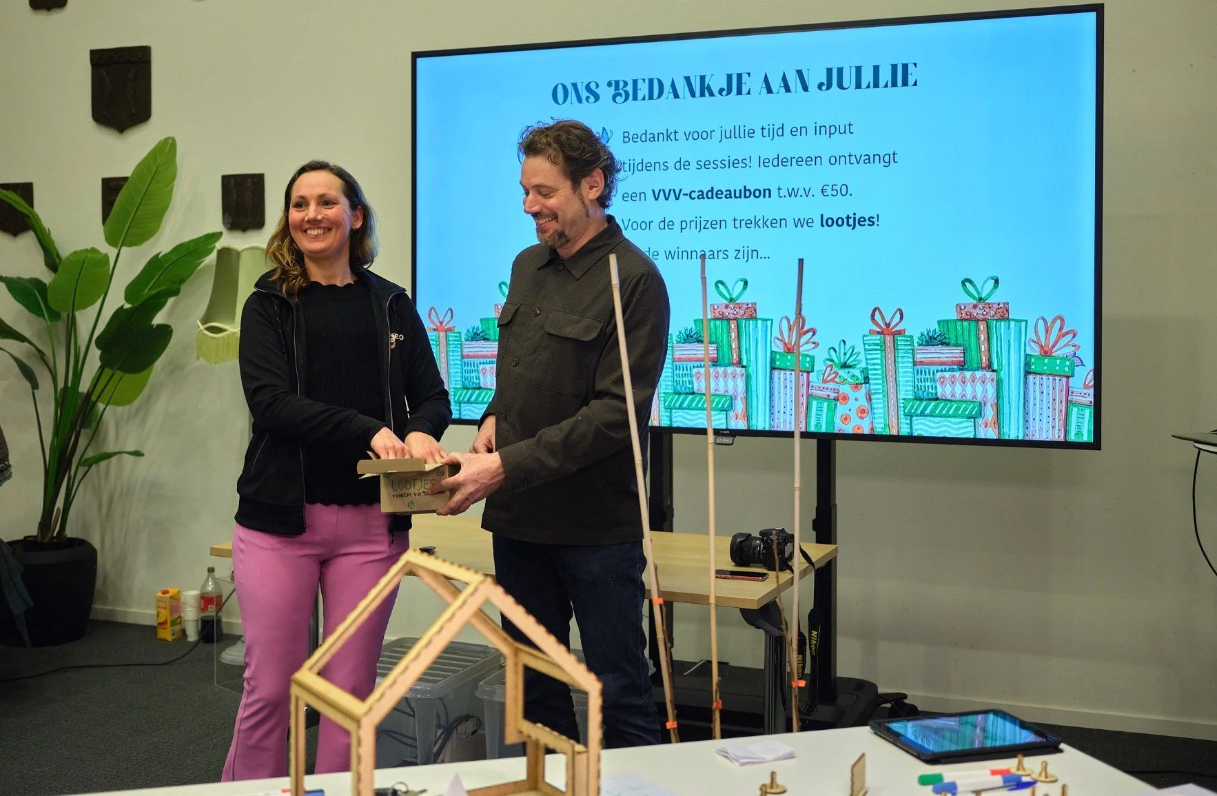 Twee mensen die een prijs of cadeaubon overhandigen tijdens een bijeenkomst of evenement, met een digitale presentatie op de achtergrond.