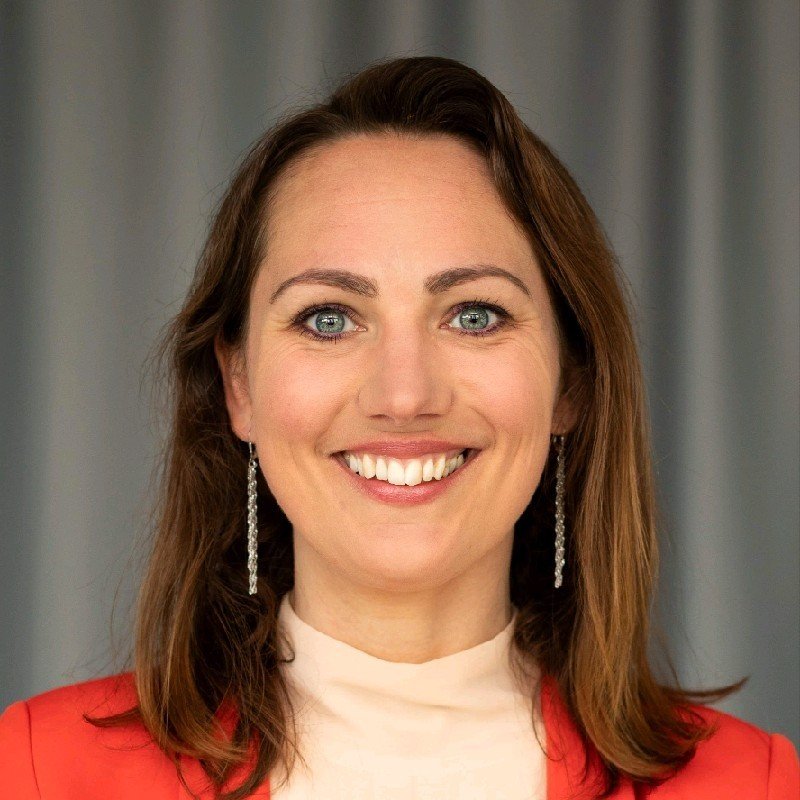 Portret van een lachende vrouw met lange bruin haar, blauwe ogen en oorbellen, gekleed in een lichte top en een rode blazer, voor een grijze achtergrond.