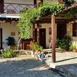 A charming brick building with a wooden door, surrounded by greenery and flowers, with a bench outside and a cobblestone pathway.