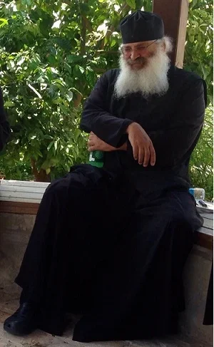 An elderly man with a white beard wearing a black robe and black head covering, sitting outdoors on a bench with greenery in the background.