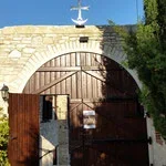 Stone archway with a wooden gate, cross on top, blue sky, and greenery in the background.