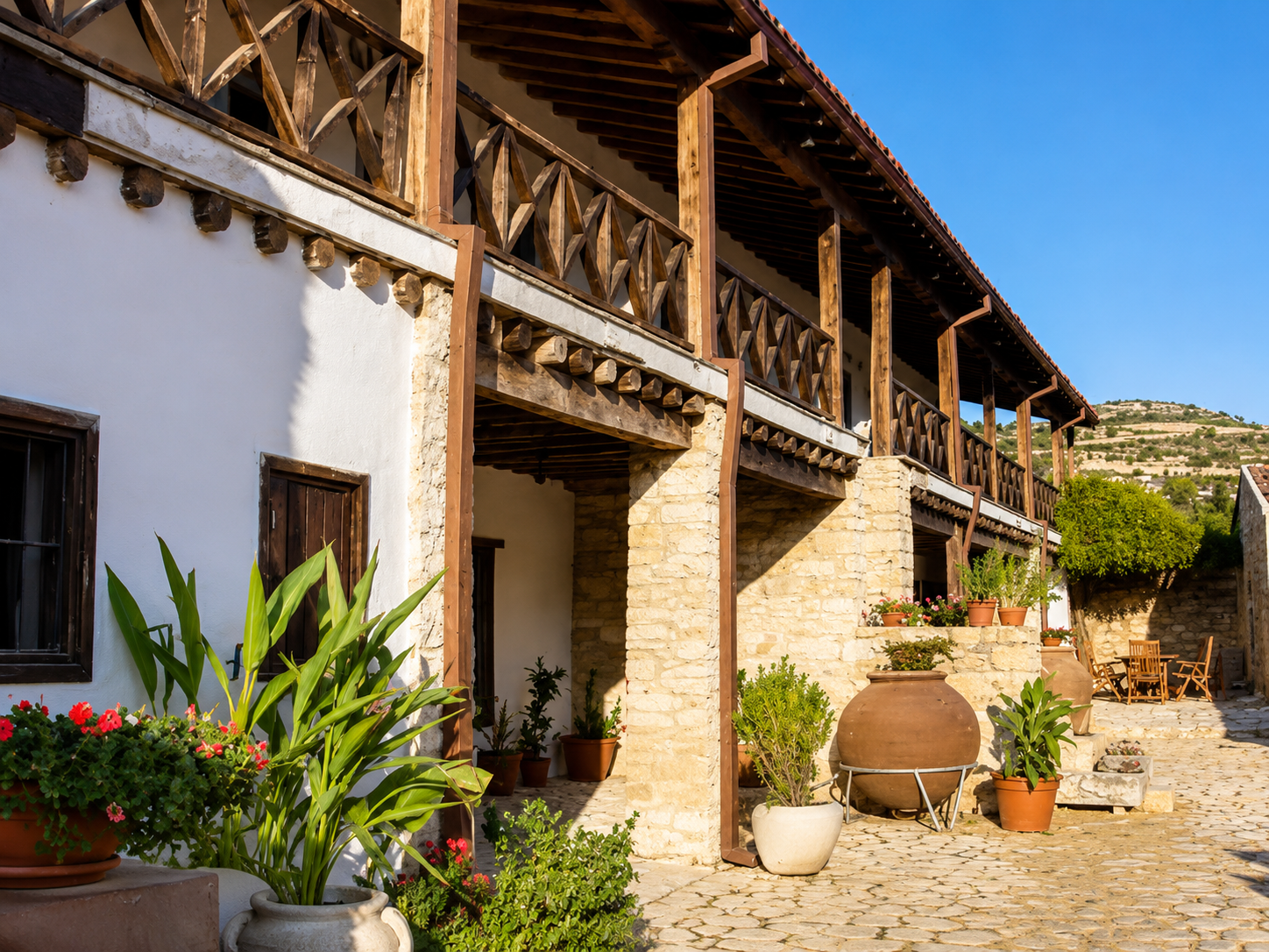 A rustic two-story house with white walls, wooden balcony, and terracotta tile roof, surrounded by potted plants and outdoor furniture, under a clear blue sky.