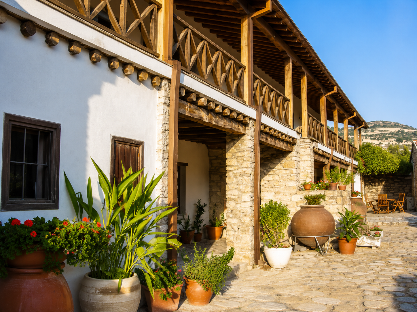 A house with a white lower level and a wooden upper balcony with a railing, surrounded by potted plants and flowers on a cobblestone patio, with a hill in the background under a clear blue sky.