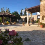 A courtyard with a stone walkway, surrounded by buildings and greenery, including pink flowers in the foreground.