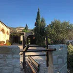 A rustic wooden gate with stone pillars, leading to a yard with trees and a yellowish building under a clear blue sky.