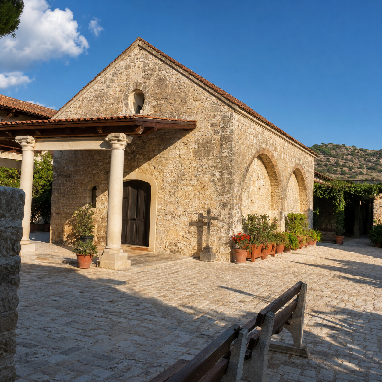 A stone church with a tiled roof, arched windows, and a small porch with columns, surrounded by potted plants on a sunny day.