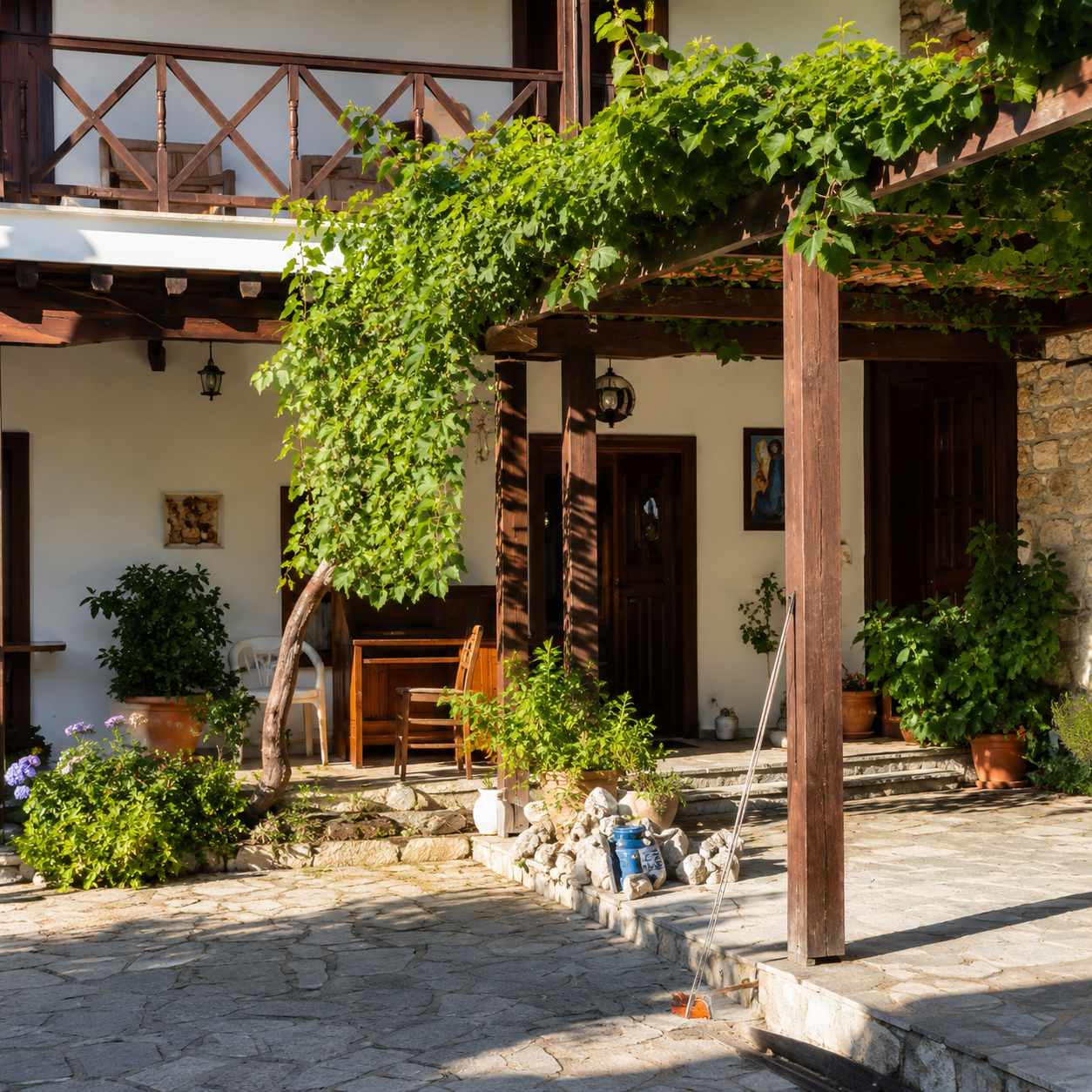 A cozy house patio with potted plants and a vine growing over a wooden pergola, with a stone paved ground, porch furniture, and decorative items like paintings and lanterns.