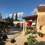 A courtyard with a stone pathway, surrounded by buildings with tiled roofs, and blooming roses in the foreground under a blue sky with clouds.