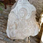 A carved stone icon of a religious figure, possibly a saint or deity, with a halo, set against a brick wall background.