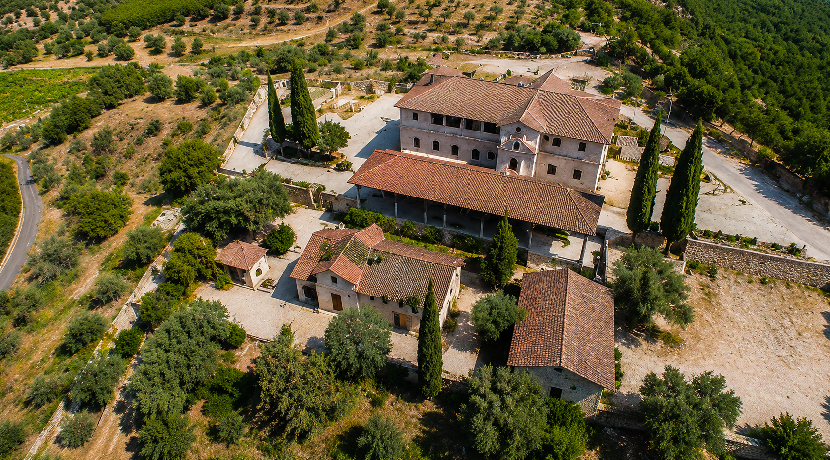 Aerial view of a historic Italian villa or monastery with multiple buildings, surrounded by trees, cypress trees, and a stone wall, located in a rural area.