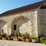 An old stone building with an arched entrance, potted plants along the base, and a red tile roof under a clear blue sky.