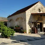 A stone building with an arched entrance and a steep roof, surrounded by greenery and a clear blue sky.