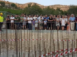 People gathered behind a fence at an outdoor event with a mountainous background.