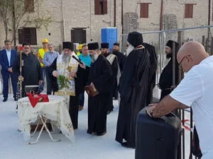 Group of people gathered outdoors around a table with a flower arrangement, part of a ceremony or event, with some dressed in formal attire, and a man with a suitcase in the foreground.