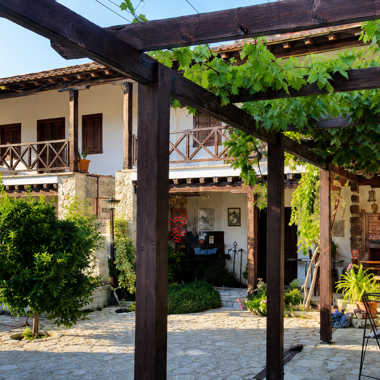 A charming outdoor courtyard of a Mediterranean-style house with stone and white plaster walls, wooden balconies, and lush green foliage, featuring a pergola with grapevines and flowering plants.