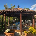 Outdoor wooden gazebo with a table and chairs, surrounded by greenery and flowers under a blue sky.