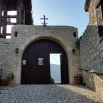Stone archway with a wooden door, situated between stone walls, and a cross on top.