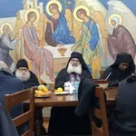 Four masked individuals sitting at a dining table with food and drinks, with a religious mural in the background.