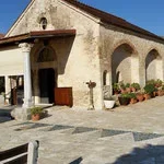 Stone building with arched doorway and windows, set on a cobblestone street with potted plants outside under a clear blue sky.