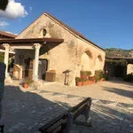 An old stone building with a clay tiled roof, surrounded by a courtyard with benches and pottery, under a clear blue sky.