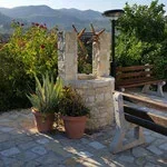 Stone well with a wooden roof and potted plants nearby, outdoor garden setting with benches and lush greenery in background.