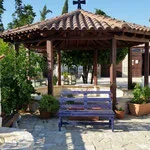 A wooden gazebo with a pointed roof, a swing hanging from the ceiling, and surrounding potted plants on a paved stone area.