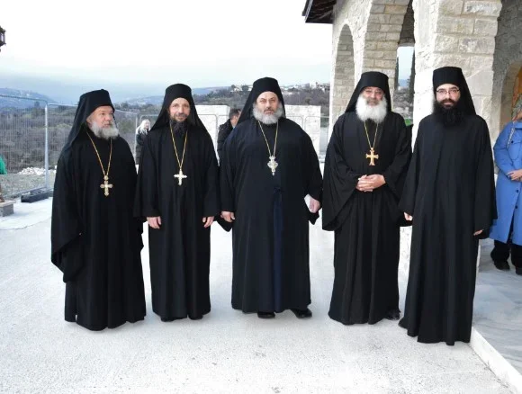 Six Orthodox Christian priests standing outdoors near a stone building, wearing black robes and crosses, with a mountainous landscape in the background.