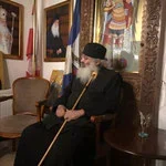 A religious leader dressed in black robes and a head covering, sitting on a chair and holding a staff in a decorated room with curtains and framed pictures.