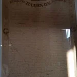 A large, weathered historical plaque or stone with engraved text, partially illuminated by sunlight, with a small window reflecting light in the background.
