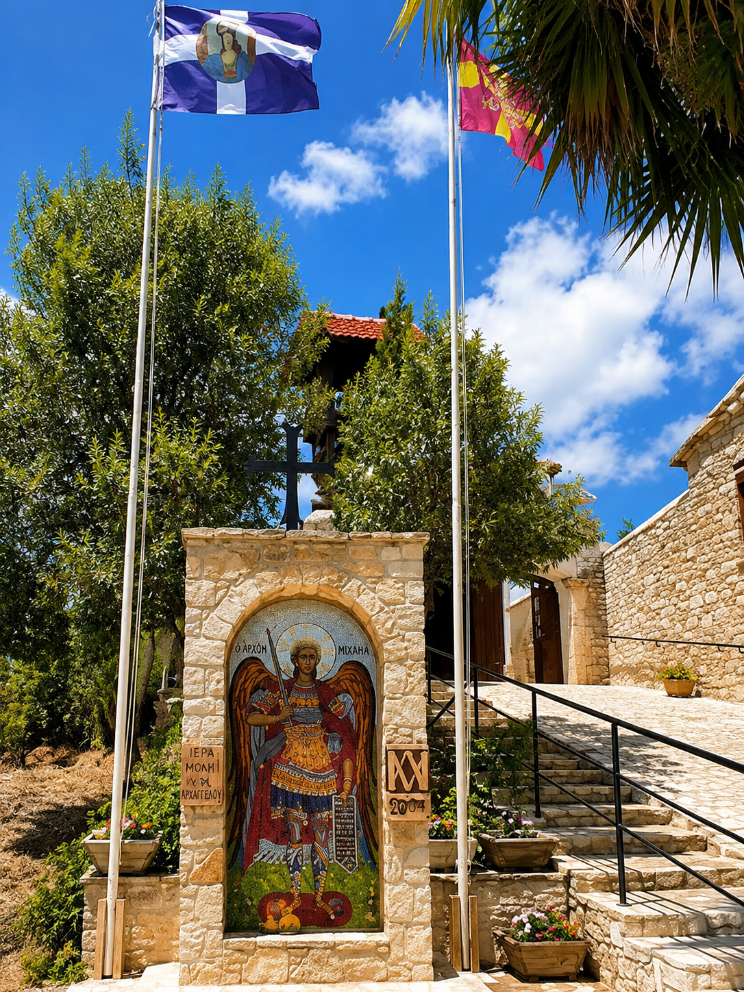 Religious shrine with icon of a saint, surrounded by flowers, stone architecture, flags, and greenery under a blue sky.