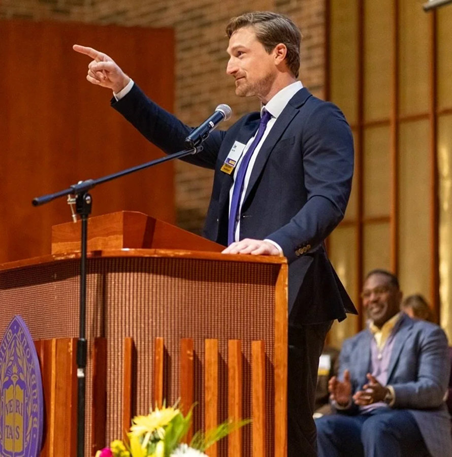 Man in a suit giving a speech at a podium during a formal event, with people clapping in the background.