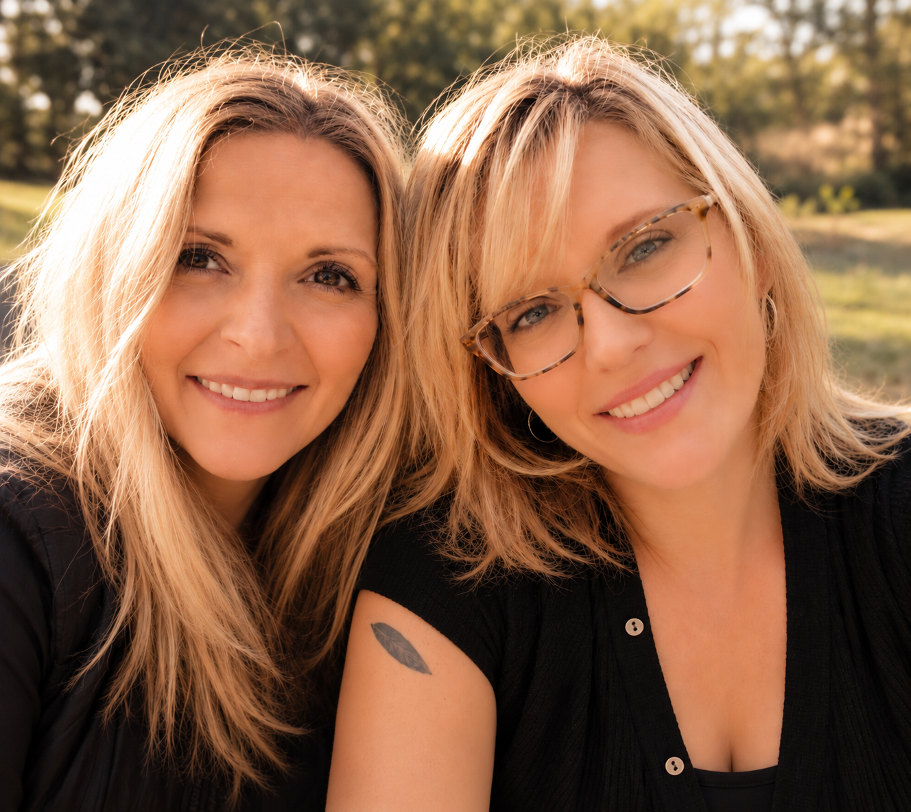 Two smiling women with blonde hair outdoors, one wearing glasses and a tattoo on her left arm.