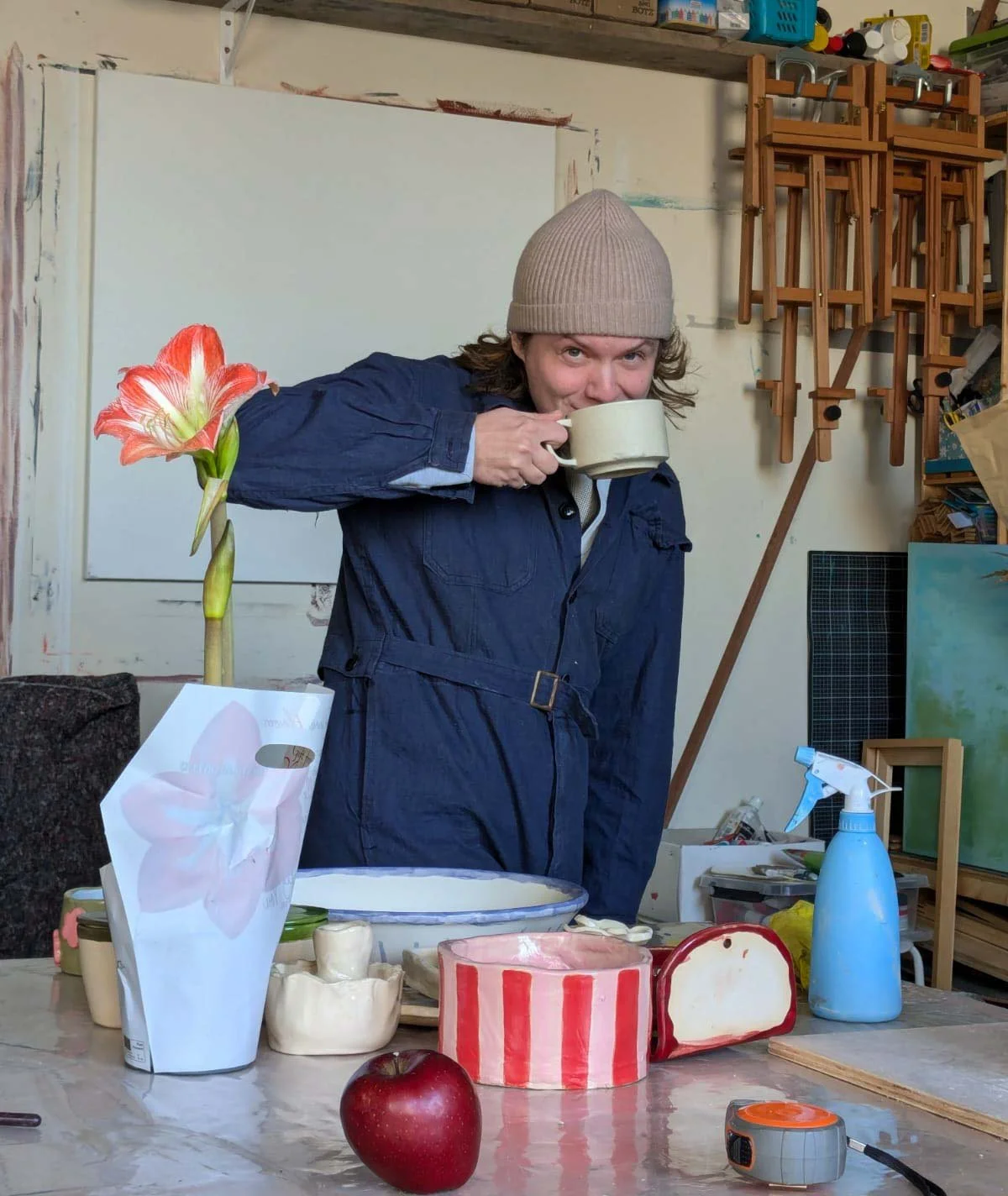 A woman with curly hair, wearing a beige knit hat and a blue jumpsuit, drinks from a large mug inside an art studio. On the table in front of her are various objects, including a tall striped cylinder with a carved face, a red apple, a white paper bag with a pink flower, a spray bottle, and some pottery.