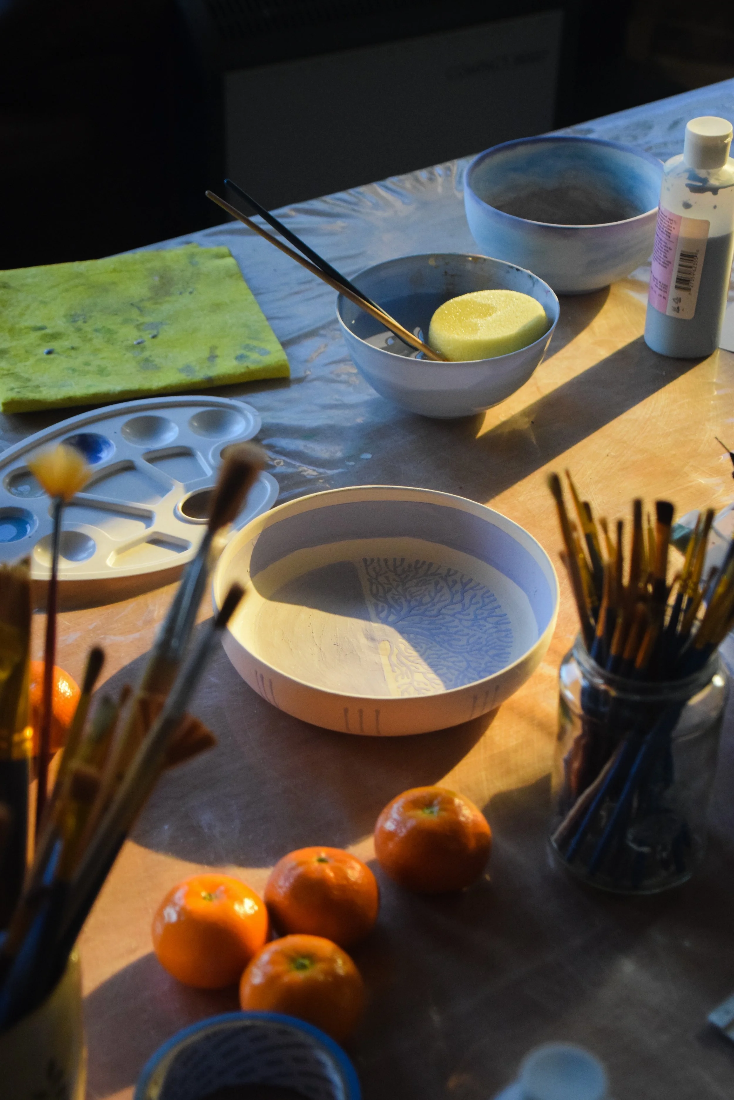 Art supplies on a table including a yellow sponge, bowls, a bottle of paint, paintbrushes, and oranges set on a wooden surface with natural light.