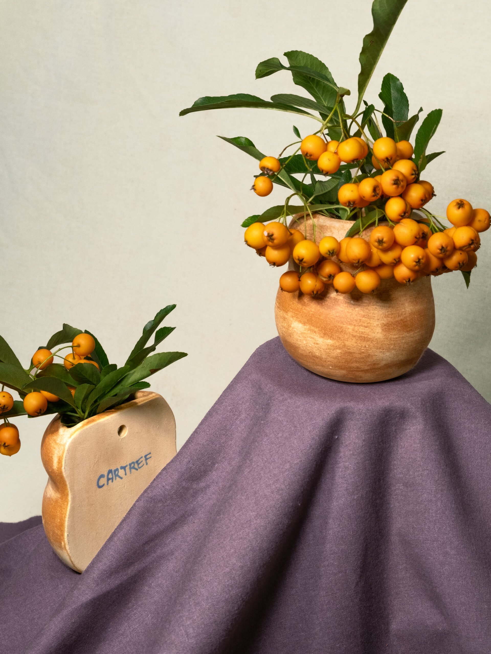 A ceramic vase with a bunch of yellow berries and green leaves, placed on a purple cloth. Another vase with similar berries and leaves is partially visible in the foreground.