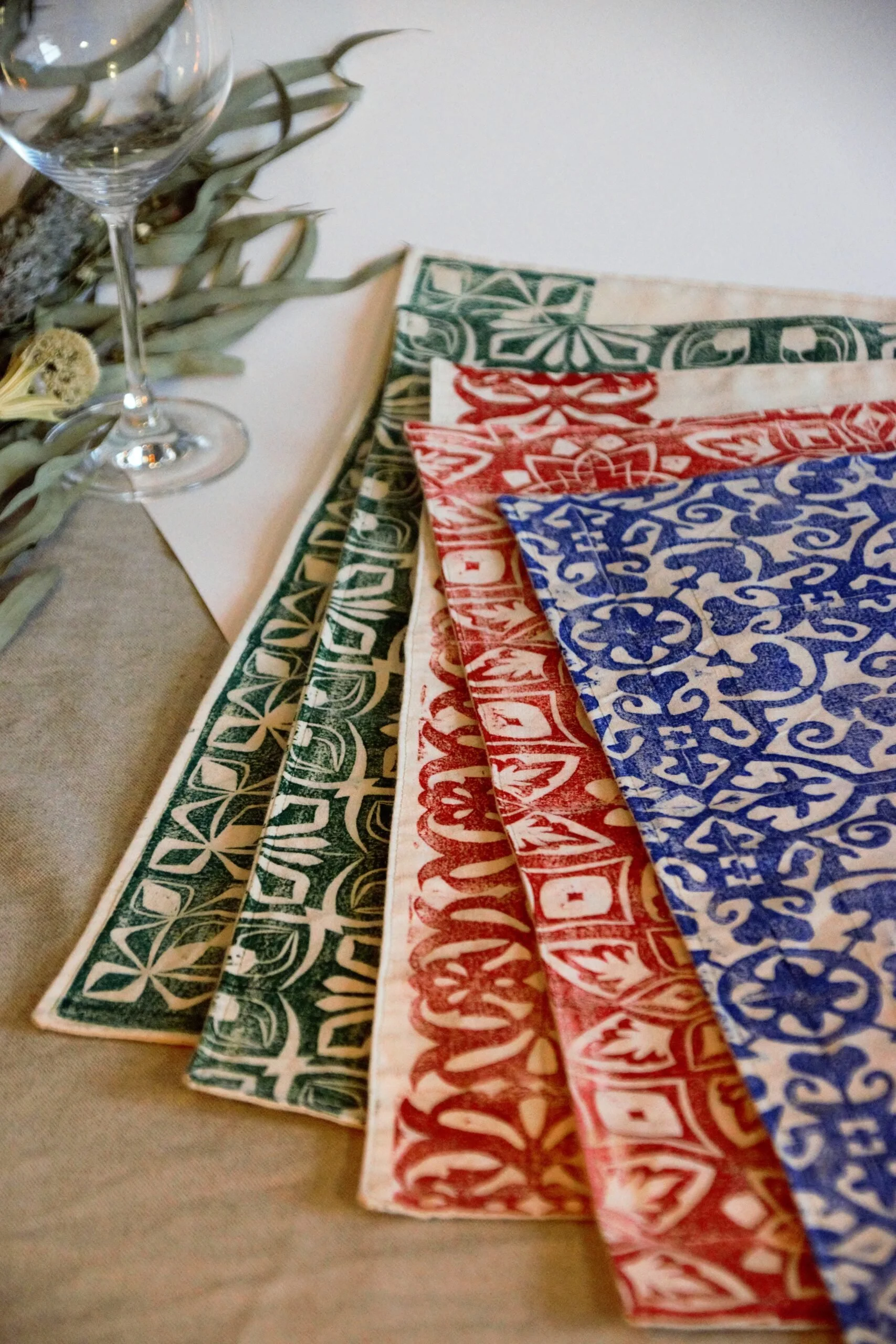 Colorful handprinted tablecloths with intricate patterns in green, red, and blue, arranged in a row on a table, next to a wine glass and dried flowers.