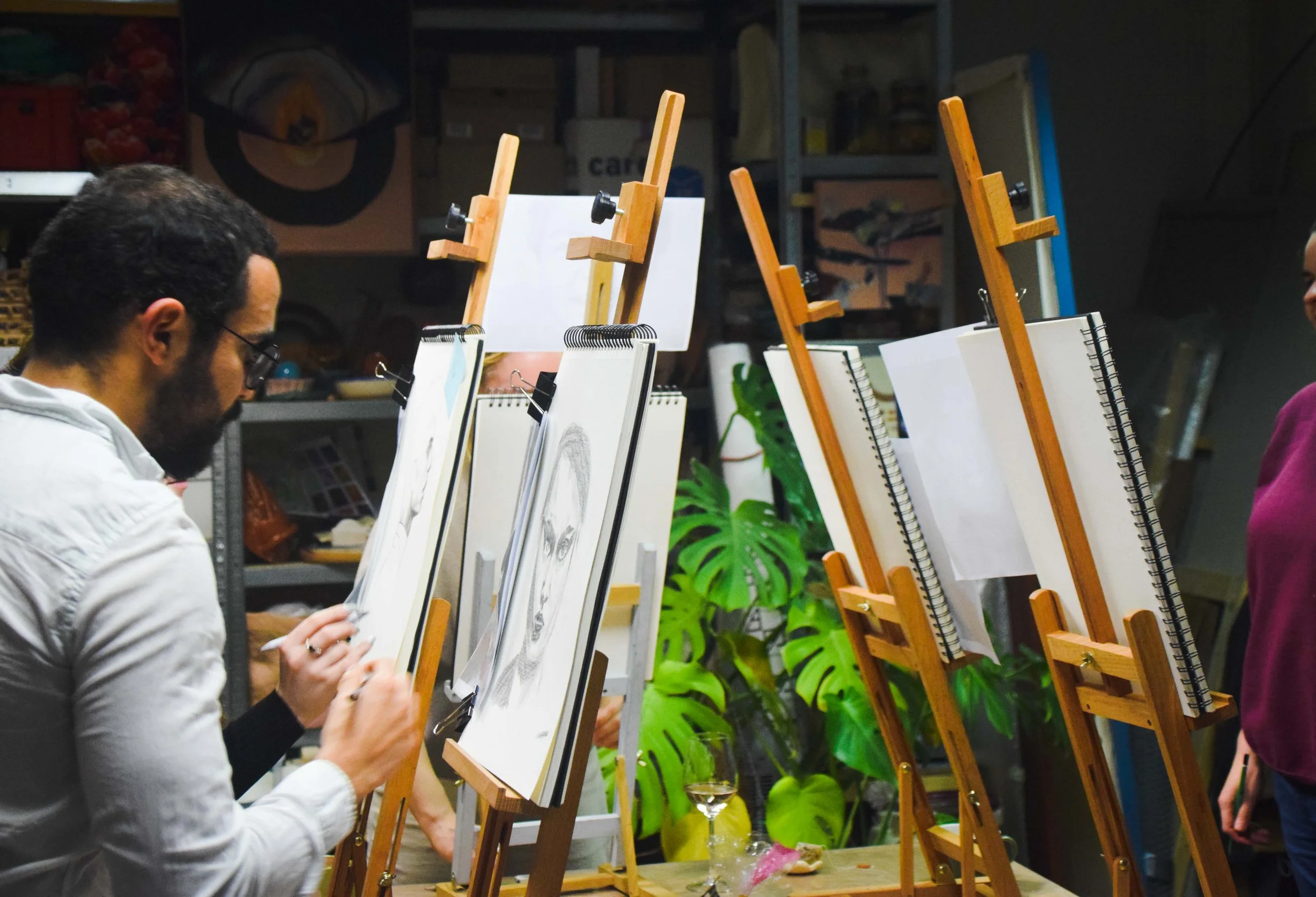 Several people are painting on canvases mounted on wooden easels in what appears to be an art studio or classroom. The background includes shelves with various supplies and artwork, and there are potted plants in the scene.