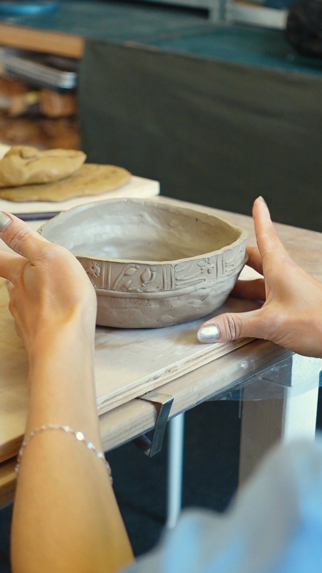 Hands shaping a decorative ceramic bowl on a wooden table in a pottery studio.