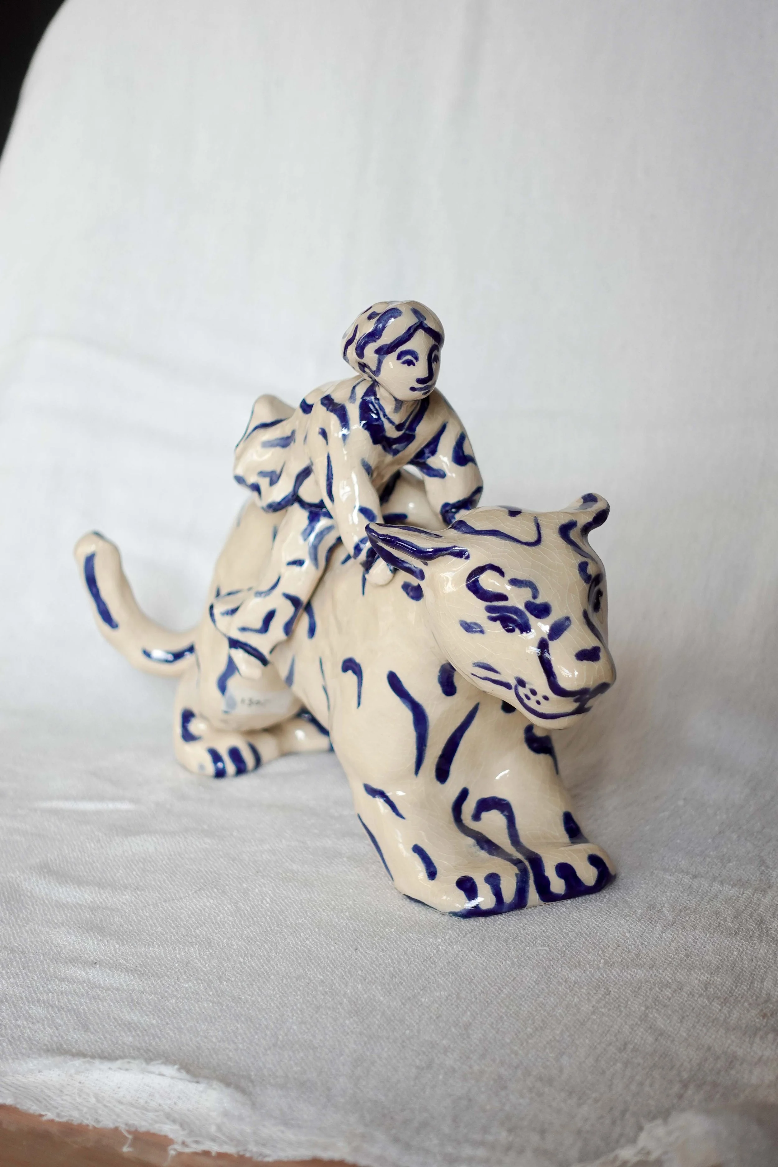 Ceramic figurine of a young boy riding a stylized cream-colored animal with blue markings, possibly a lion, on a light-colored fabric surface.