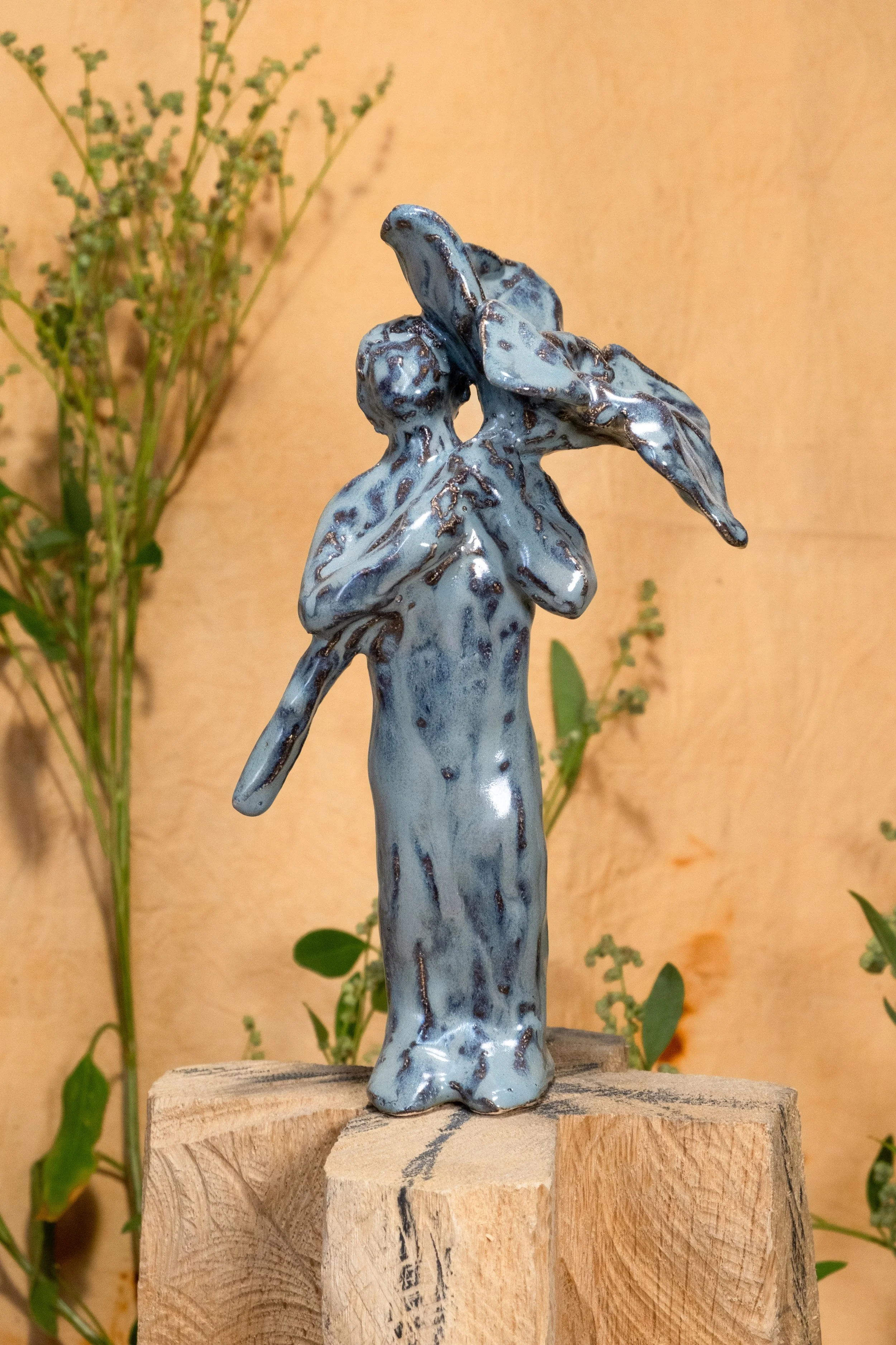 A glazed ceramic statue of a woman with a flower, standing on a wooden block with a background of green plants.