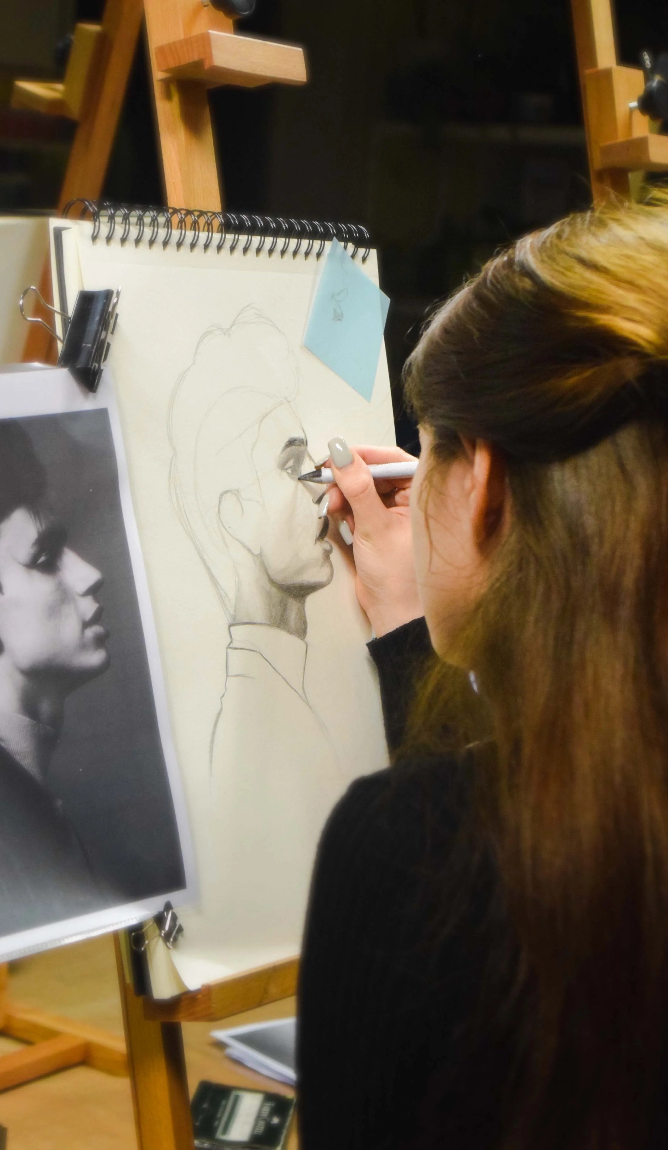 A woman with brown hair is drawing a detailed pencil sketch of a woman in profile on a spiral-bound paper. There is a black and white photo of the same profile on a nearby sheet.