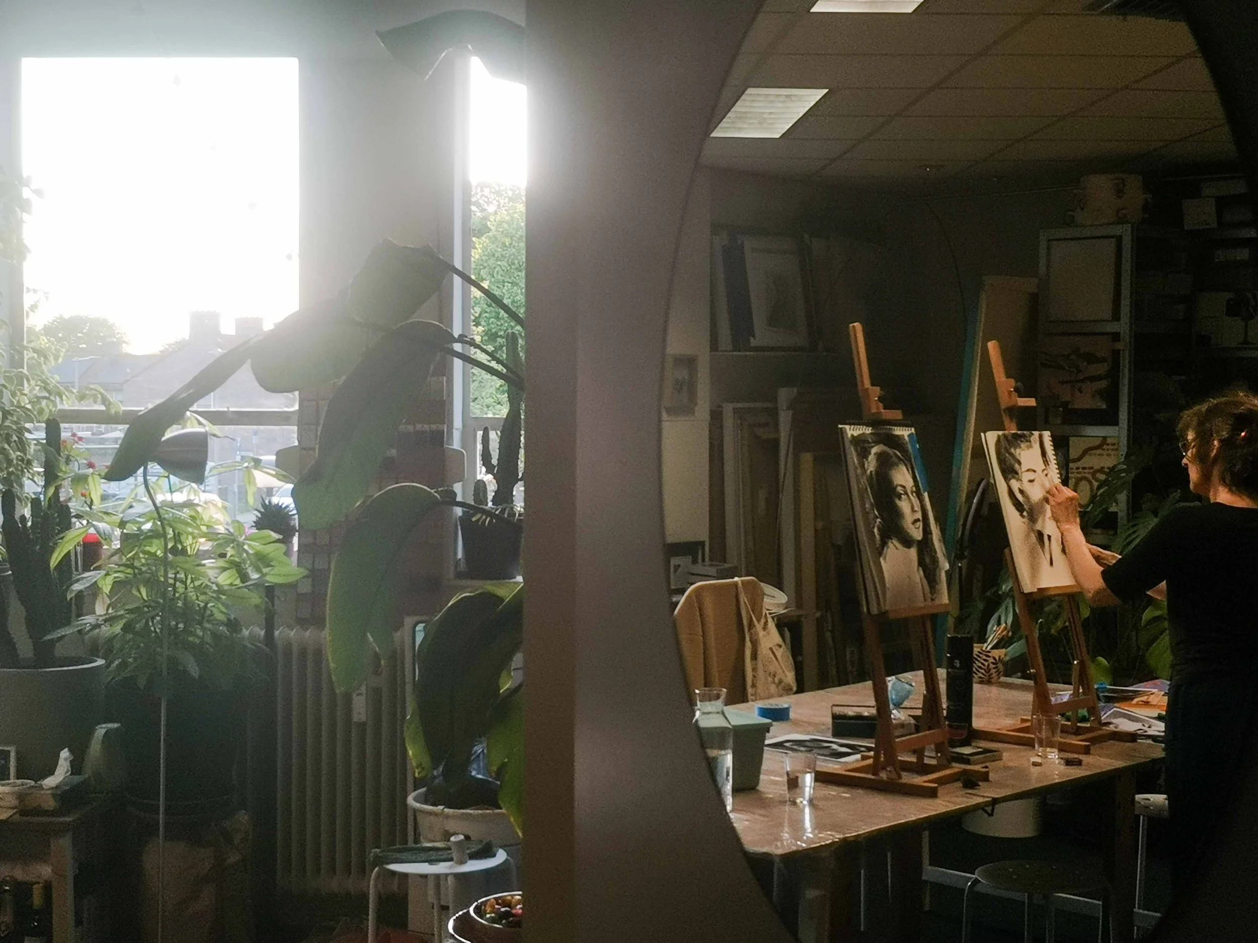 Cartref's creative art studio in Arnhem with an easel and portraits of women, plants in pots near a bright window, and various art supplies on the table, with a person working on an artwork.