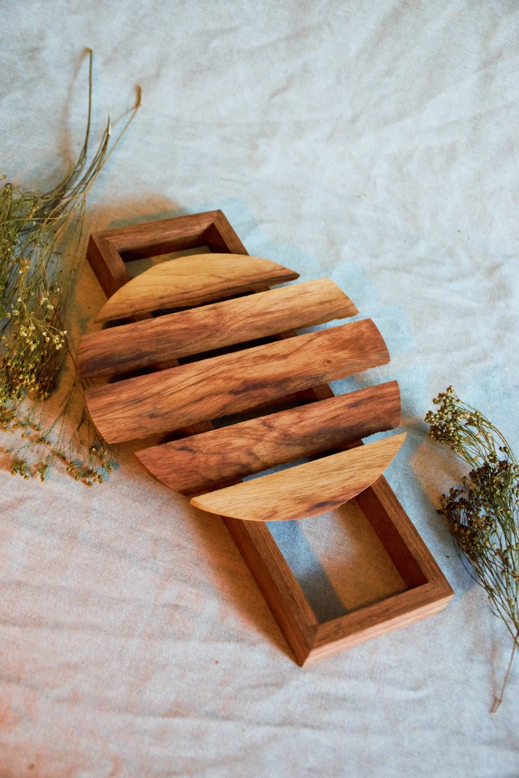 Wooden bathroom or kitchen soap dish with a slatted design, on a fabric surface, with some small dried flowers nearby.