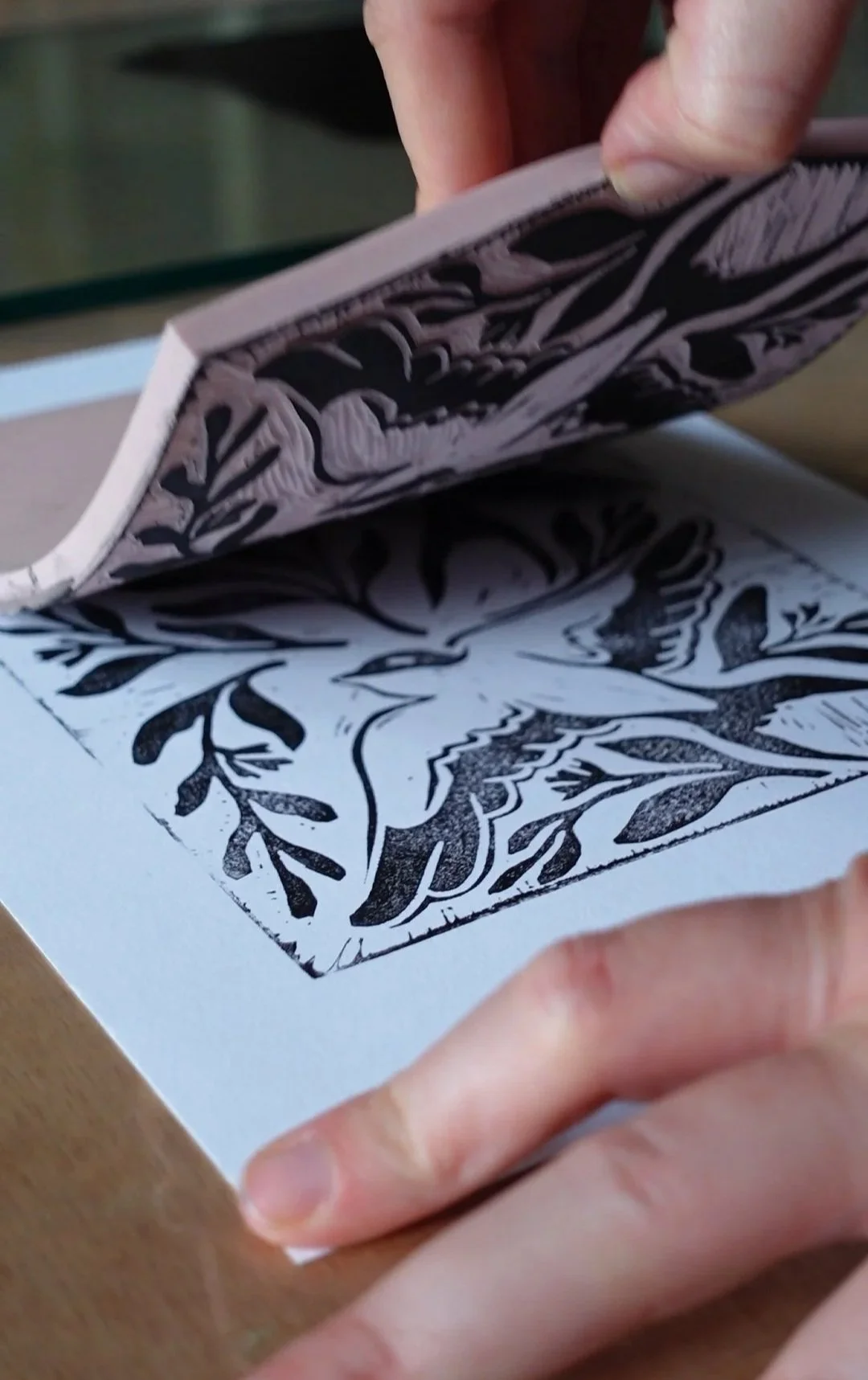 Close-up of hands lifting a printed linocut block to reveal a carved black-and-white floral pattern on paper.