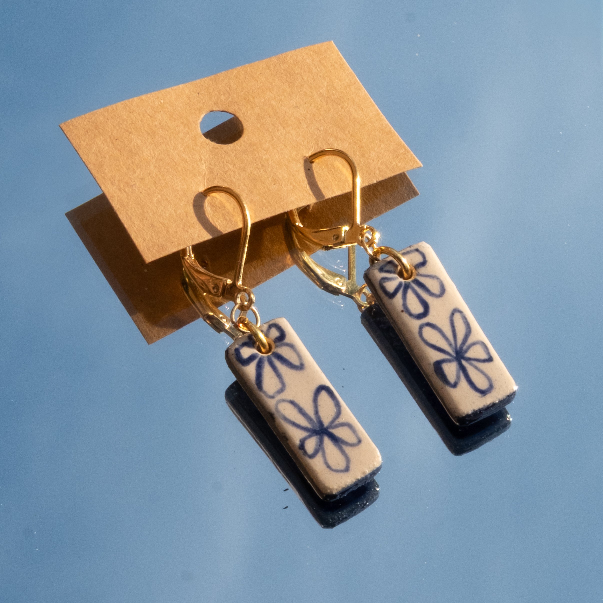 Close-up of two ceramic earrings hanging on a gold-colored hook, attached to a small piece of cardboard with a hole, against a blue background.