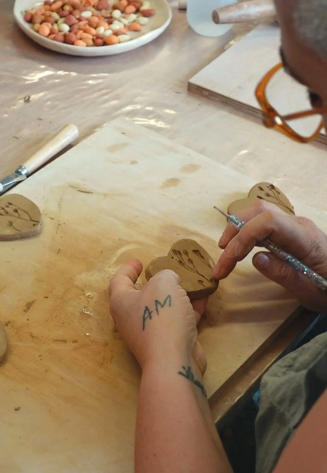 Person carving a design into a heart-shaped piece of wood with a small carving tool. The person's left hand has the word 'AM' tattooed on the wrist. In the background, there is a plate with small, round beans or seeds.
