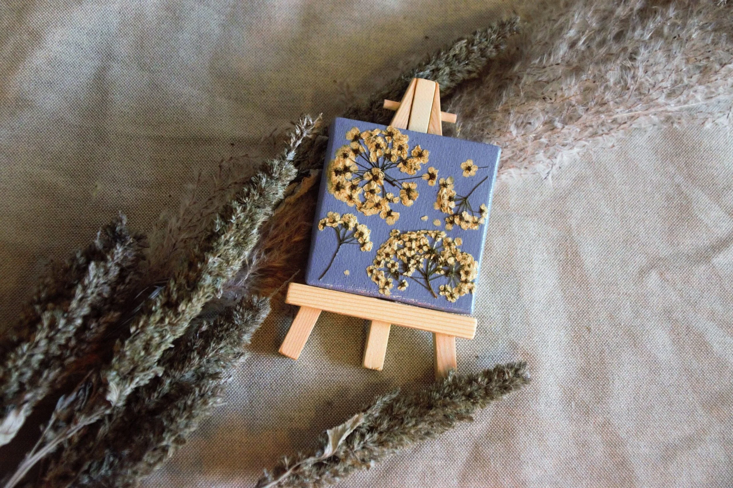 A small painted canvas on a miniature easel, decorated with natural dried yellow flowers and green stems, resting on a textured beige fabric background, surrounded by dried fluffy pampas grass.