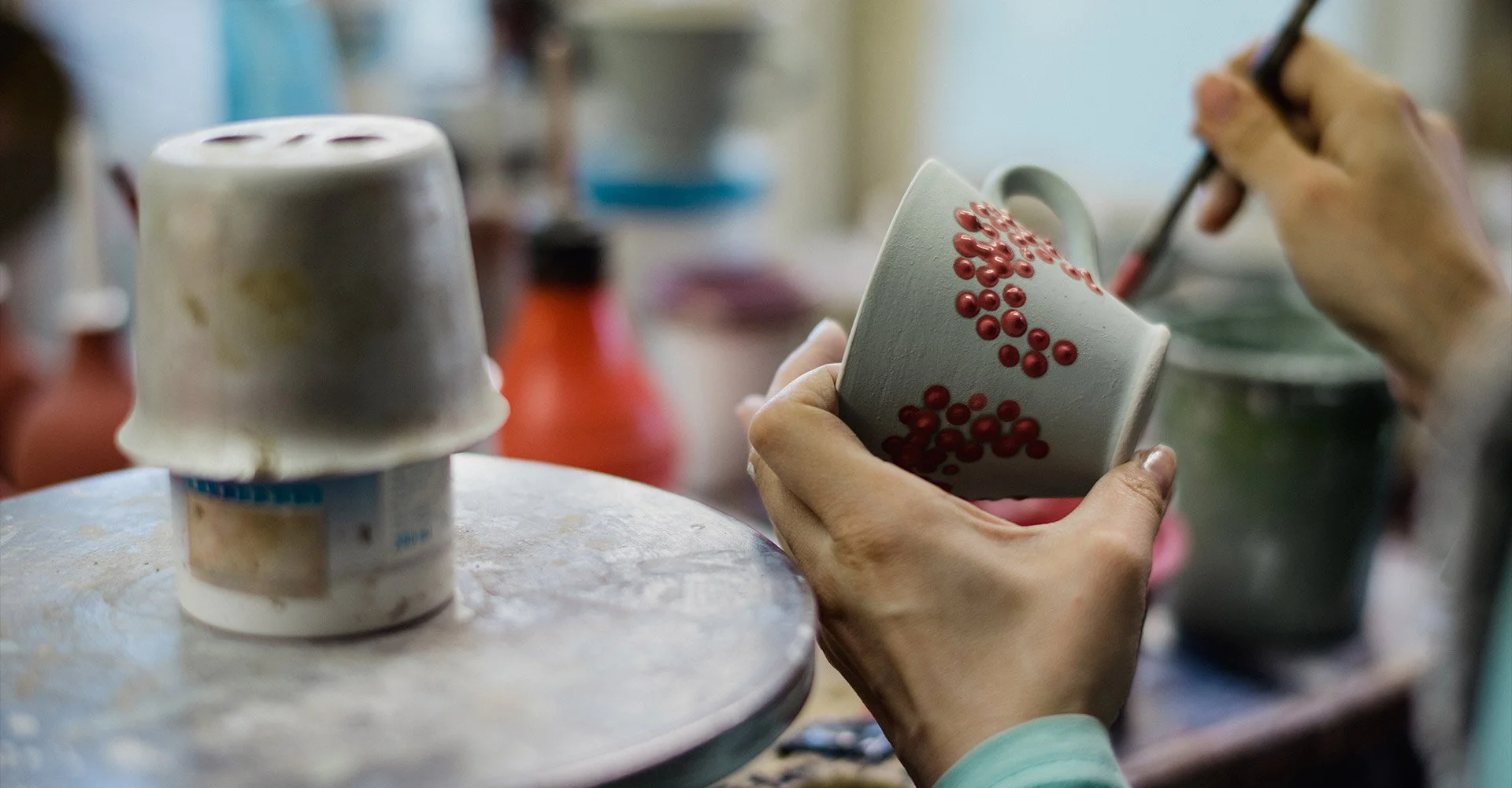 A person painting red dots onto a white mug, with another mug and various art supplies visible in the background. In Arnhem ceramic studio atelier.