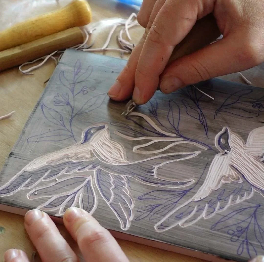 Person carving a floral design into a lino sheet with chisels and gouges, with outlines sketched in purple ink and ballpoint pen. Created in Arnhem Cartref creative art studio.