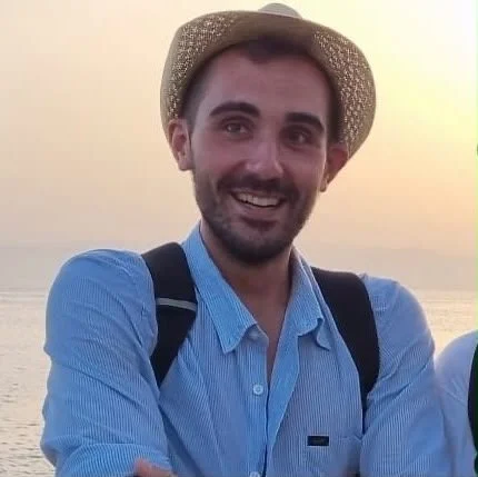 Uomo sorridente con cappello di paglia e camicia azzurra, sul mare al tramonto.